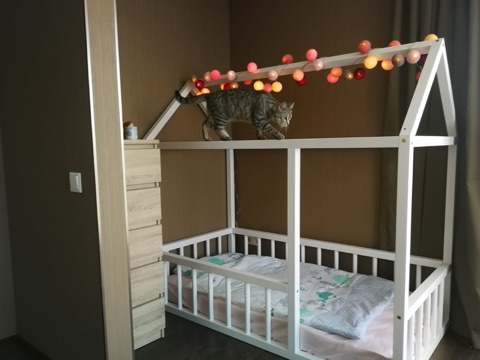 Montessori gultiņa bēŗniem ar standarta augstuma bortiņiem un apaļām redelēm. Bērnu mājiņgulta ar skursteni un brusu, kas veido durtiņas. Bērnu gultiņa baltā krāsā.